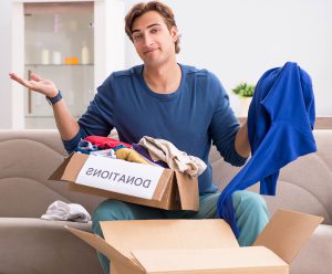 A man holding a sweater in one hand, with both hands up in the air. A box on his lap is full of clothes and is labeled "donations."