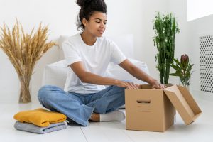 A young woman sits on the floor and folds clothing into a box to donate it to charity